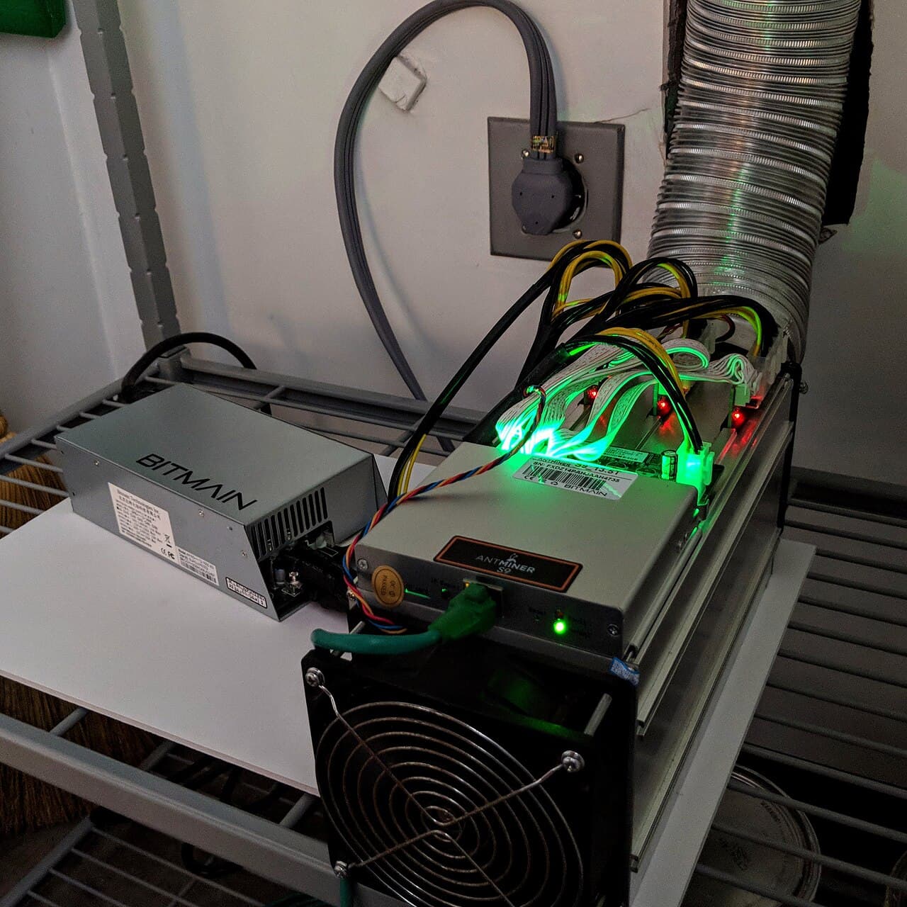 A Bitmain Antminer S9 next to a Bitmain power supply unit, with an exhaust duct behind it
